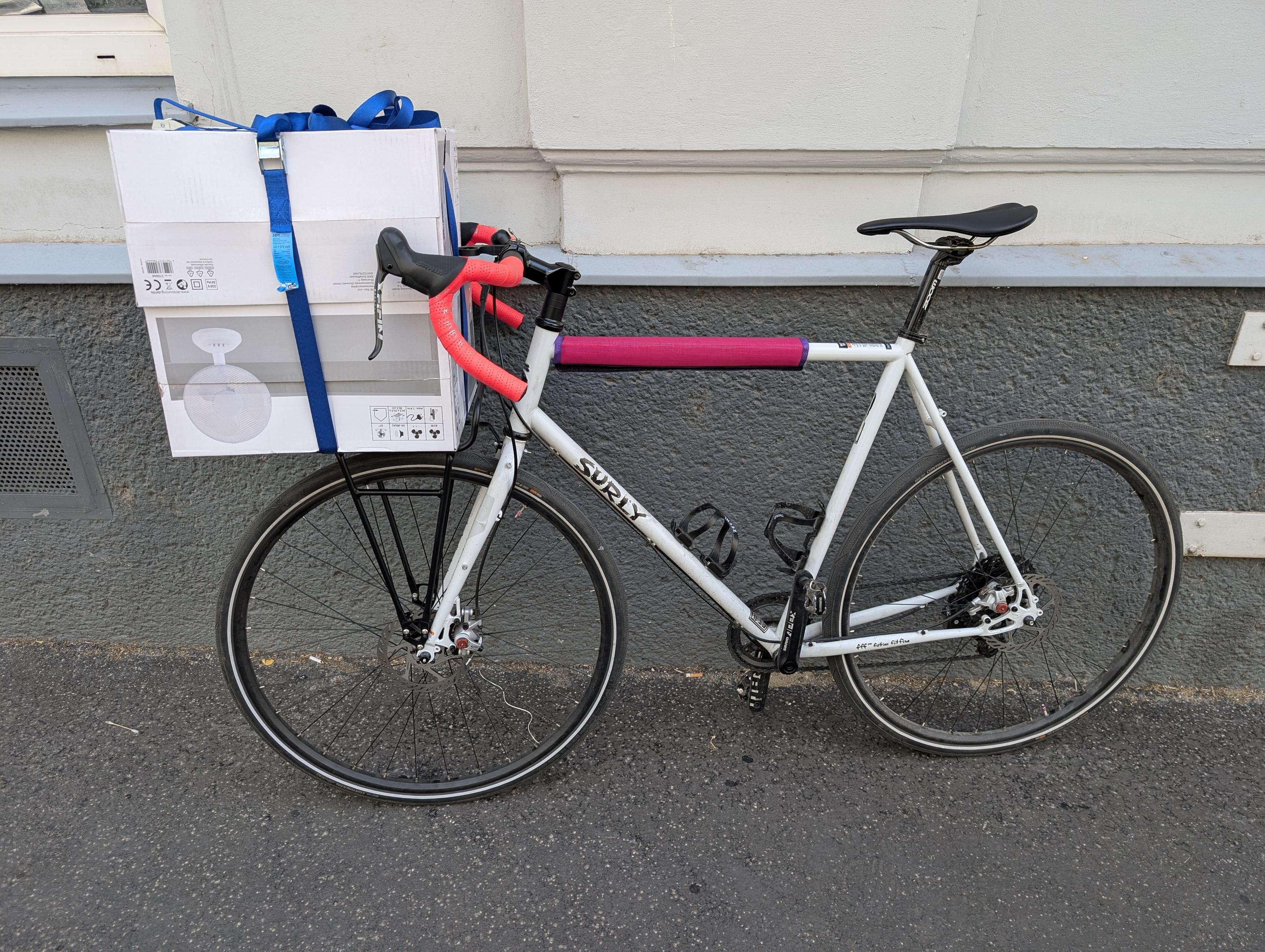 Foto eines weißen Rennrads mit rosa Lenkerbank und pink-lila Oberrohrschutz aus Stoff. Vorne zwischen den Bremshebeln sind auf einem Vorder-Gepäcksträger zwei Kartons mit jeweils einem Tischventilator gepackt, die mit zwei blauen Zurrgurten befestigt sind.
